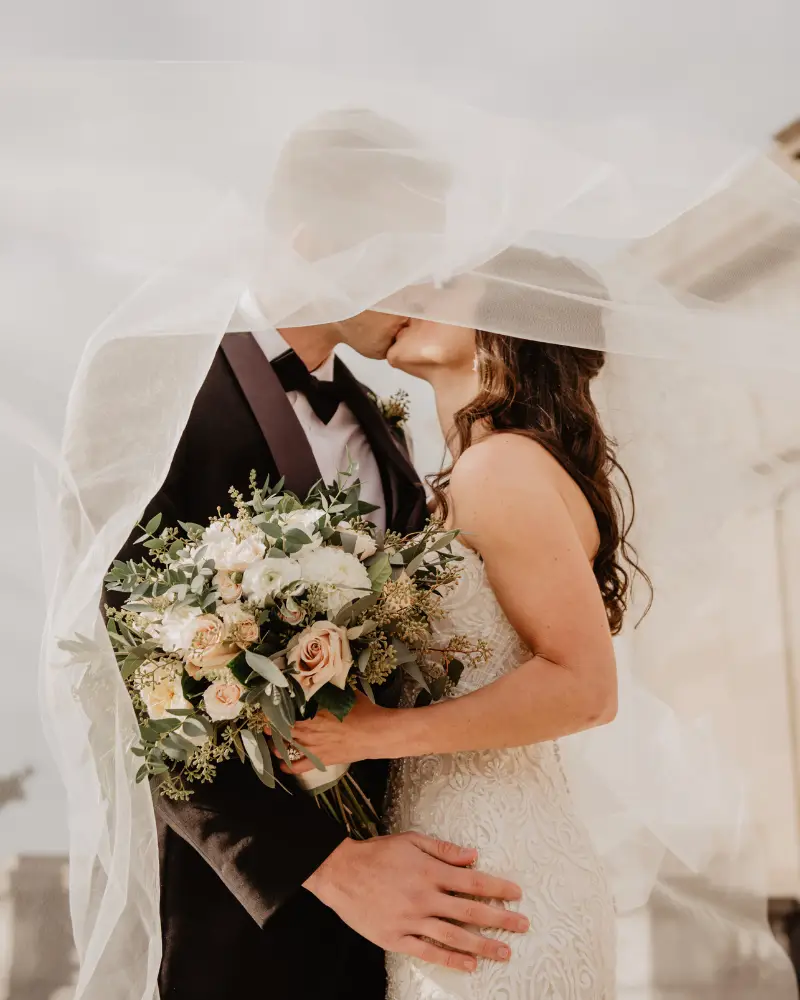 Zelf je bruiloft plannen, maar met een beetje hulp van mij (Femke) op de achtergrond. Ik ben geen typische weddingplanner, maar help jou terug naar rust en overzicht om weer heerlijk in jullie bruidsbubble te komen en relaxt je eigen bruiloft te organiseren. Foto: bruidspaar onder sluier met bruidboeket duidelijk zichtbaar.